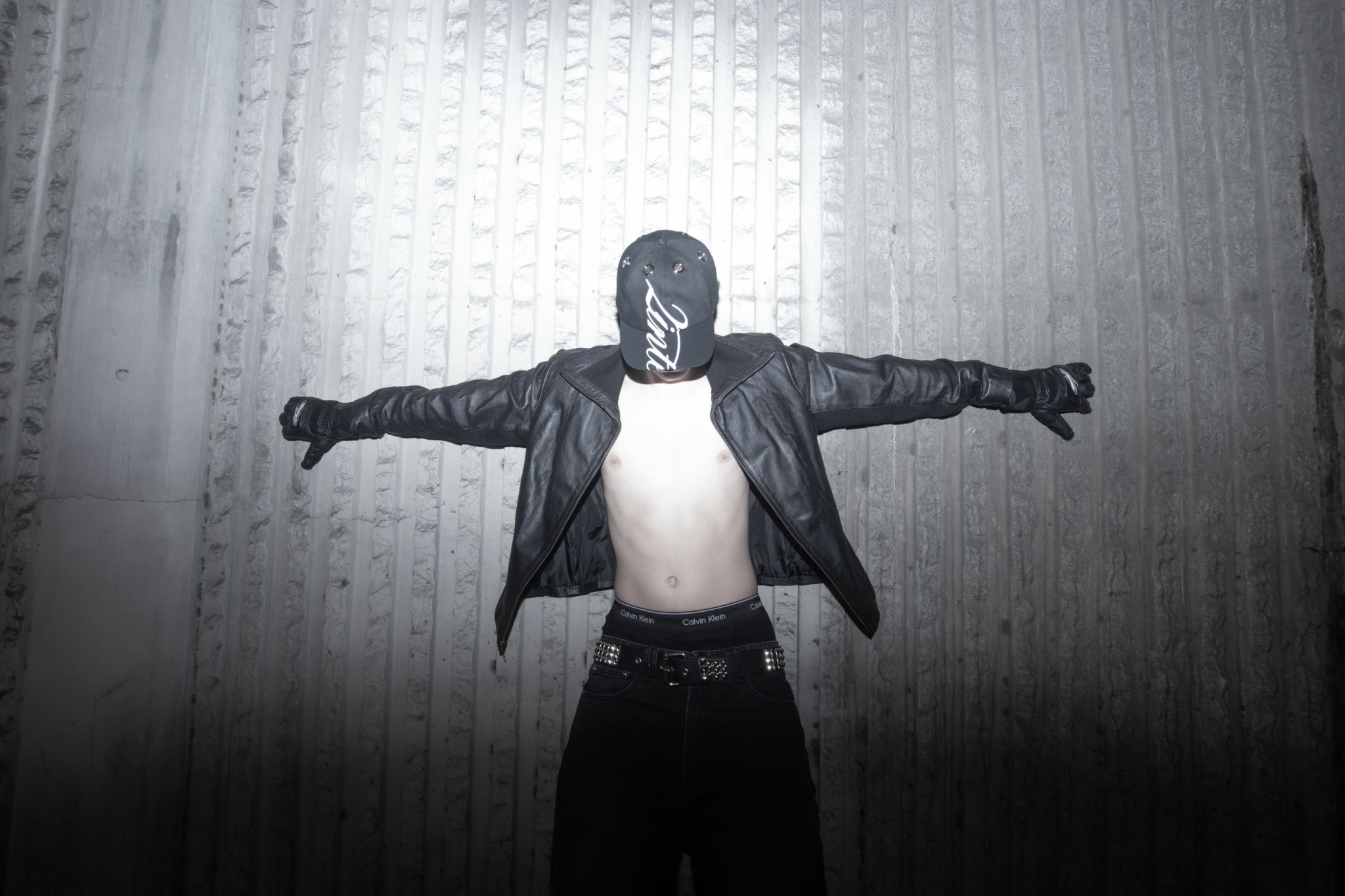 Person with arms outstretched, wearing a mask, leather jacket, and gloves, standing against a textured wall.