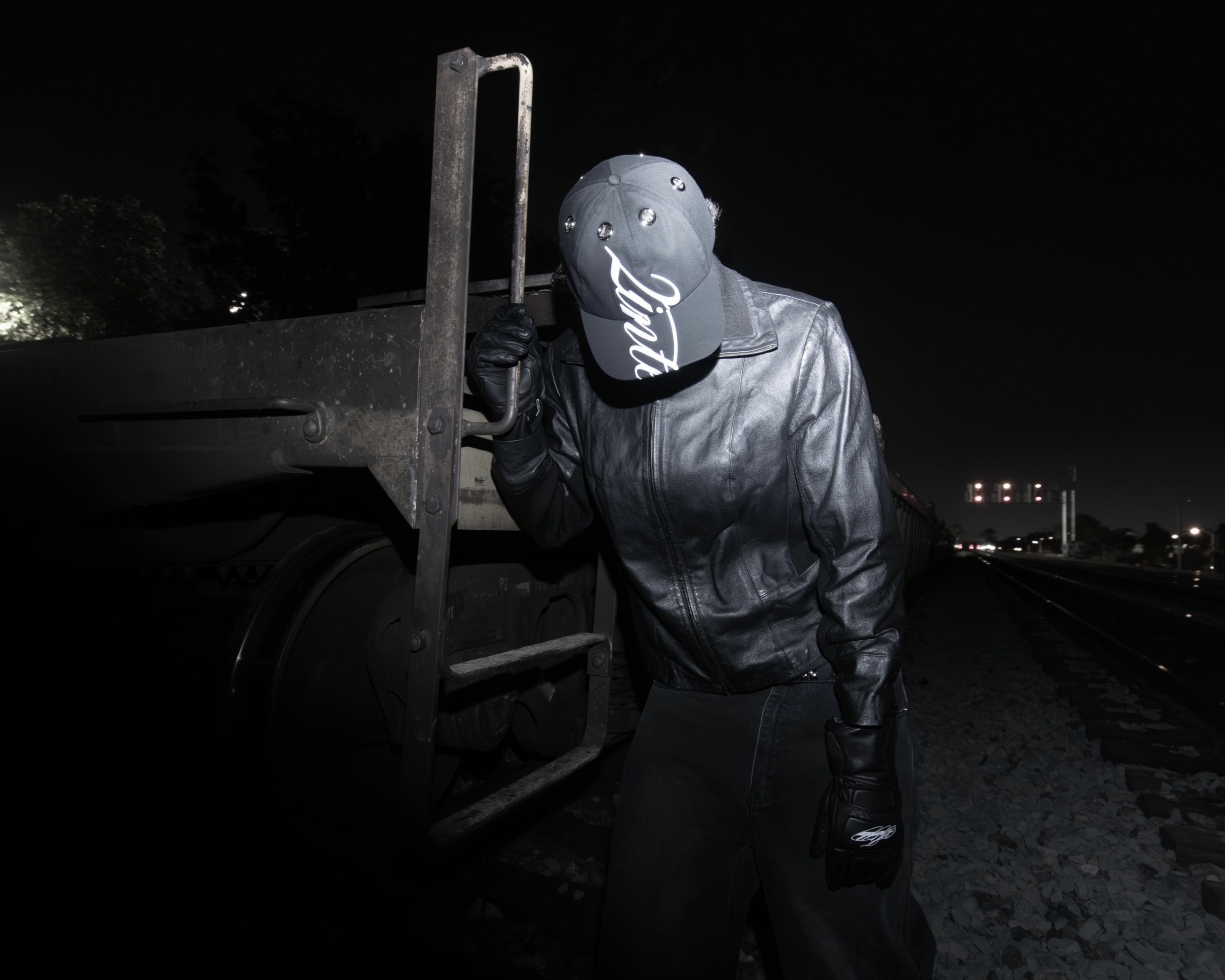 Person wearing a cap and leather jacket leaning against a truck at night, with a dark sky and distant city lights.