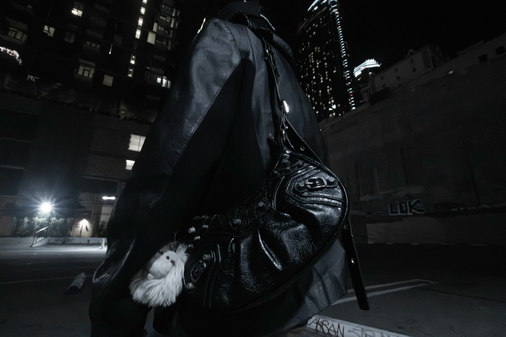Person wearing a leather jacket and carrying a black bag with a white fluffy keychain, cityscape at night.