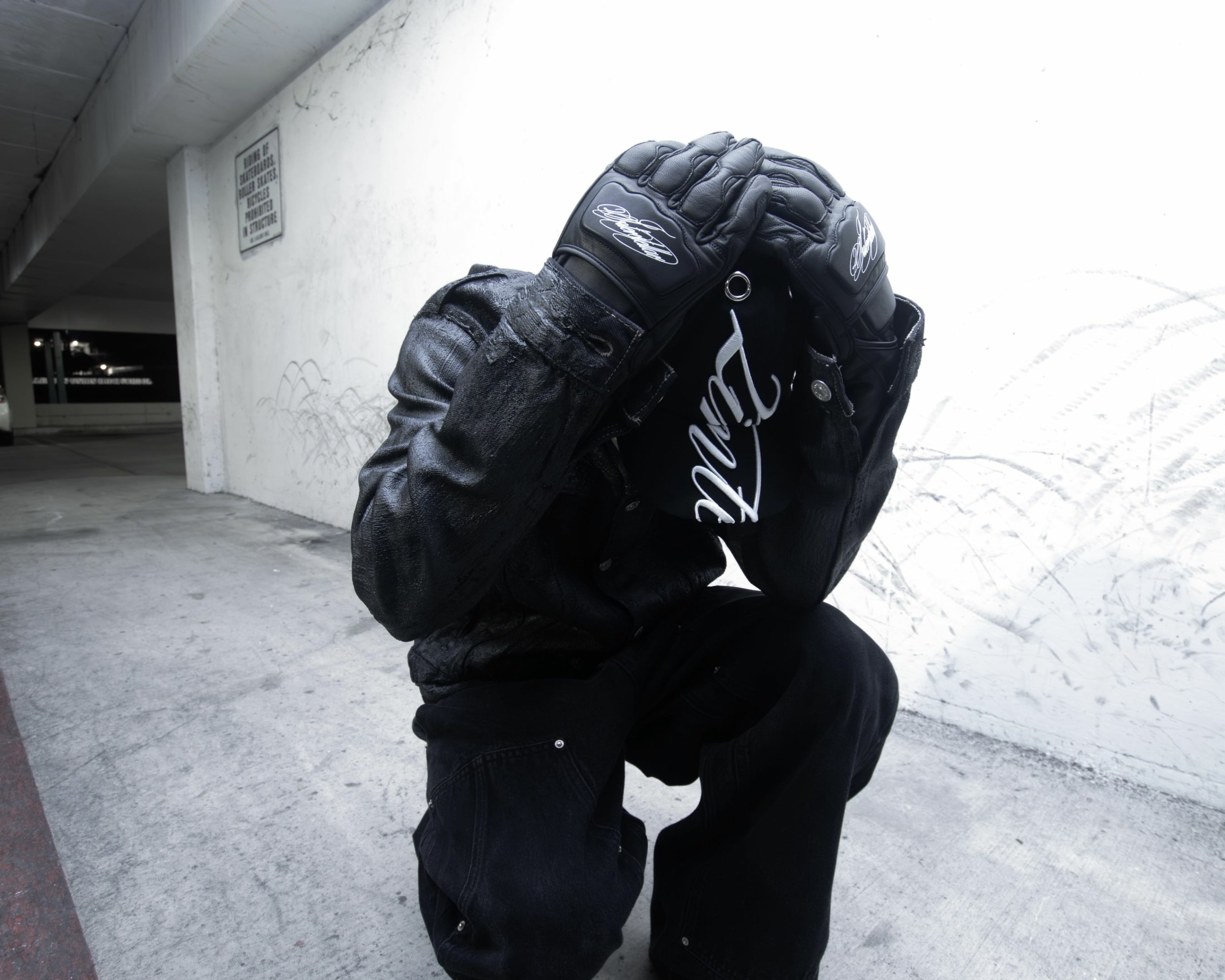 Person crouching with hands covering face, wearing black gloves and dark clothing, in an urban setting.