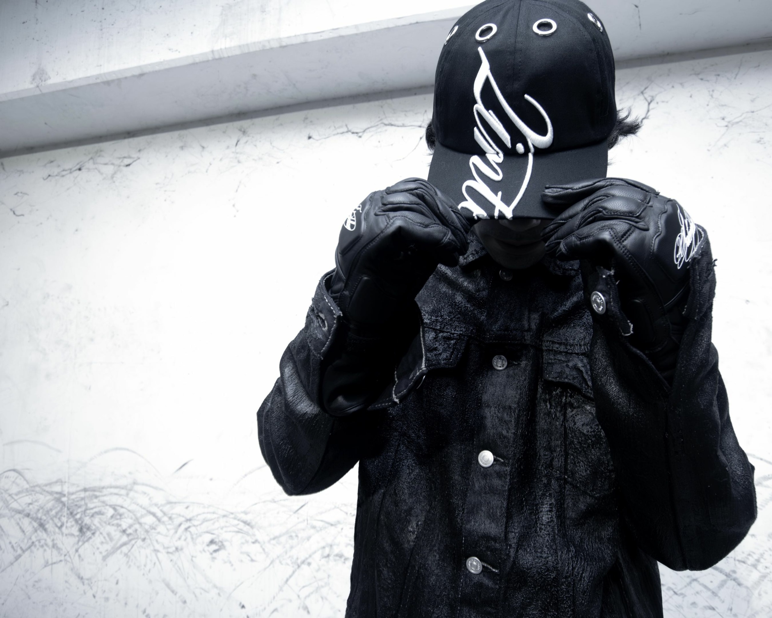 Person wearing a black hoodie and jacket, with a face covering, standing against a white wall.