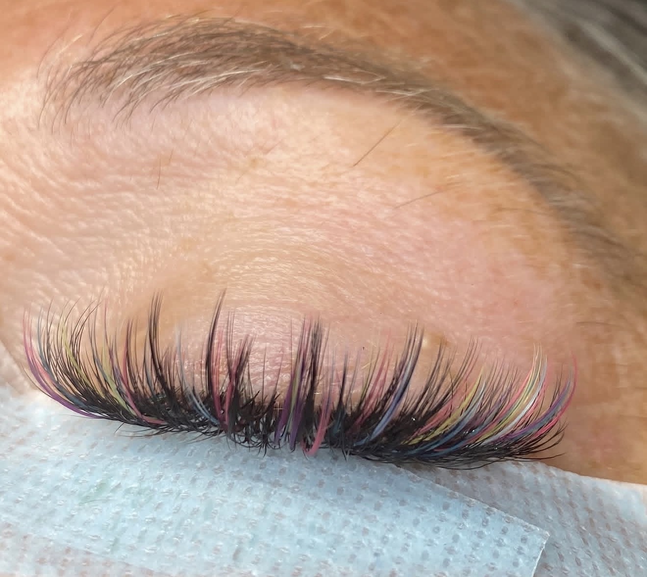 Close-up of a person's closed eye with colorful eyelash extensions in pink, blue, and yellow hues.