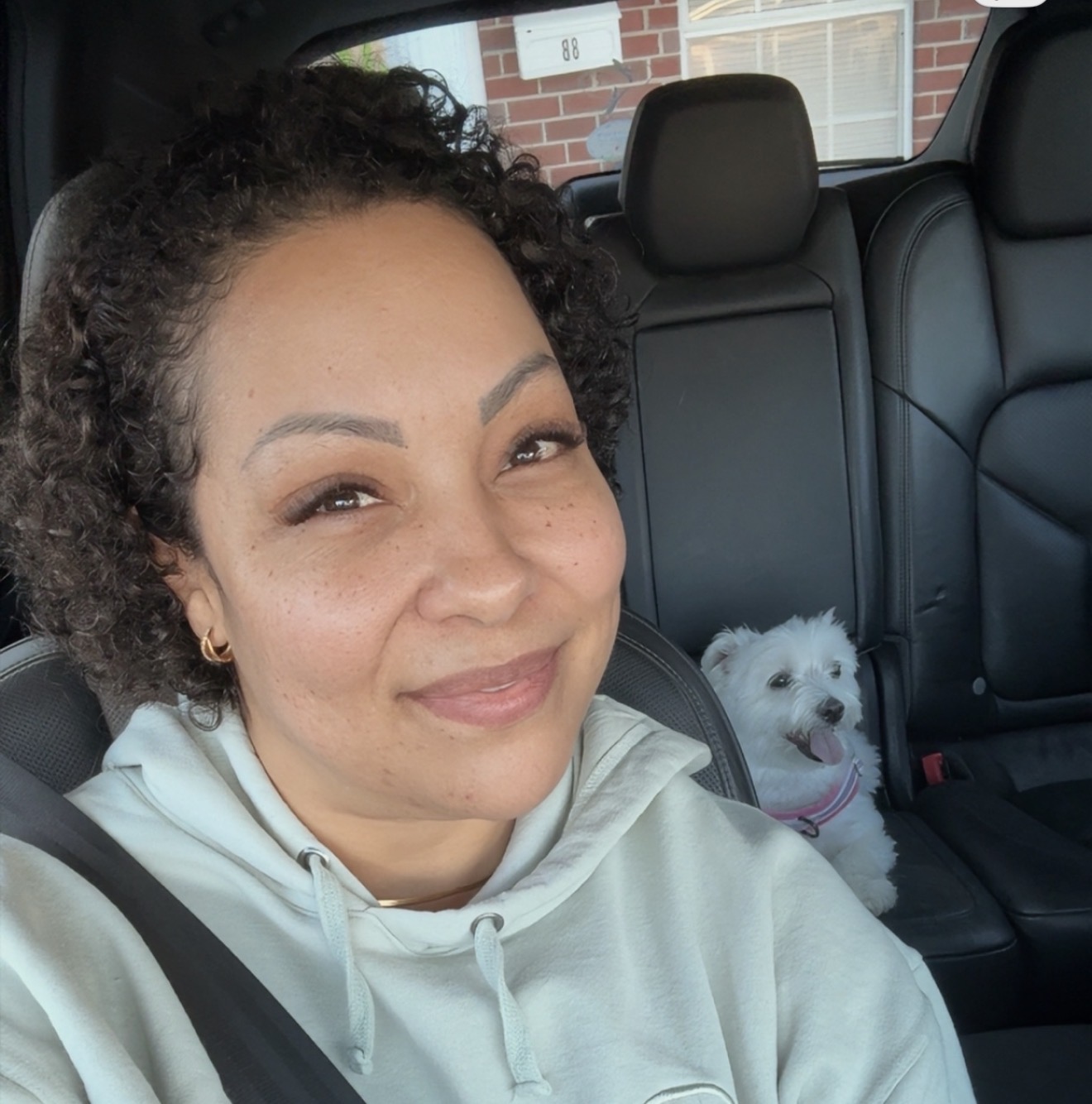 Woman with curly hair smiling in car, small white dog in back seat, brick building visible outside window.