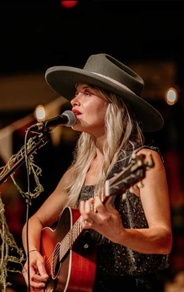 Woman singing into microphone while playing guitar, wearing a wide-brimmed hat, in a dimly lit setting.