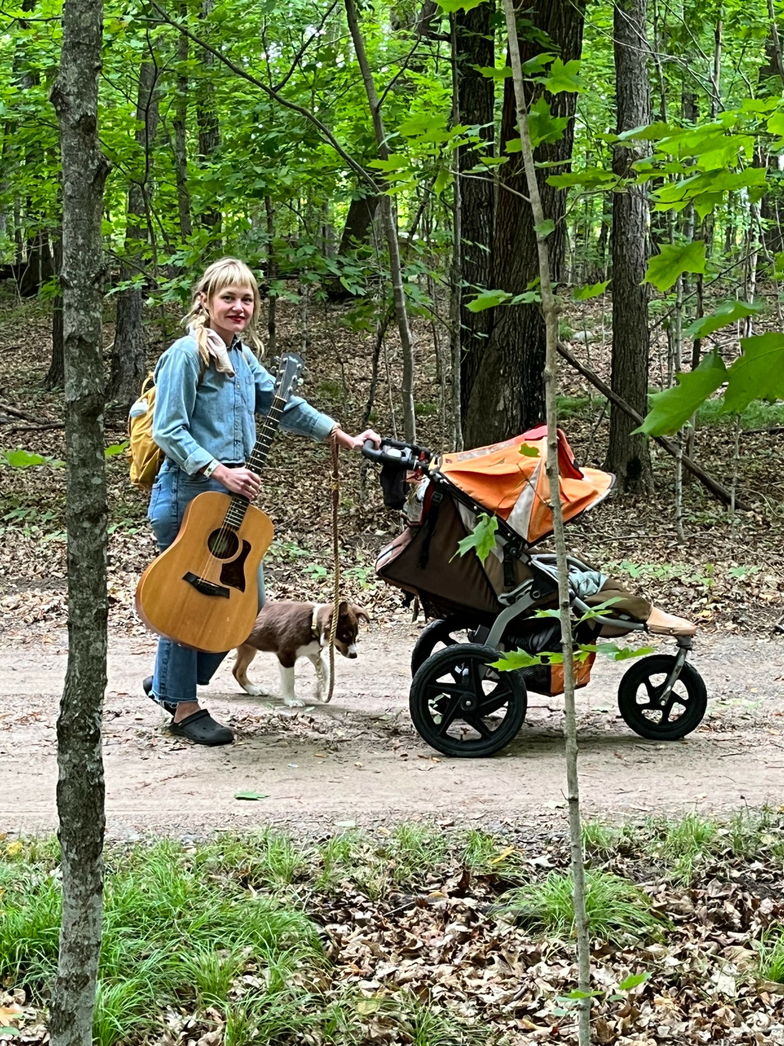 Girl with guitar and backpack in forest, holding a leash with a small dog, next to a stroller with an orange cover.