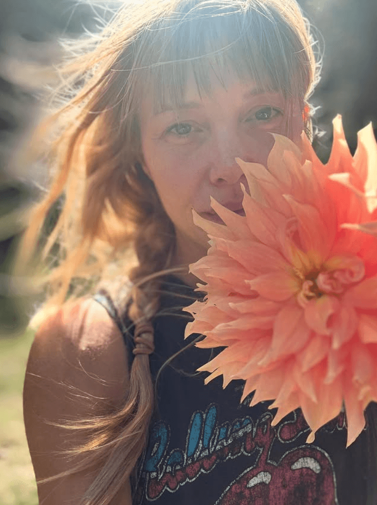 Woman with light brown hair and bangs holding a large pink flower close to her face outdoors.