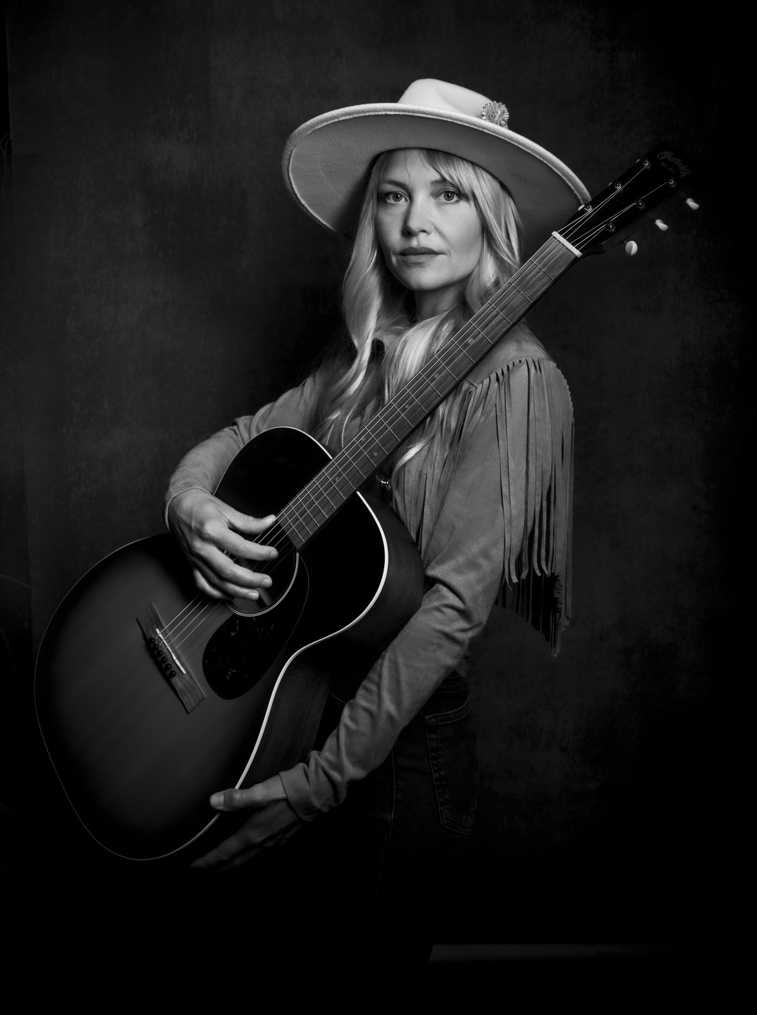 Woman wearing a wide-brimmed hat holding an acoustic guitar against a dark background.