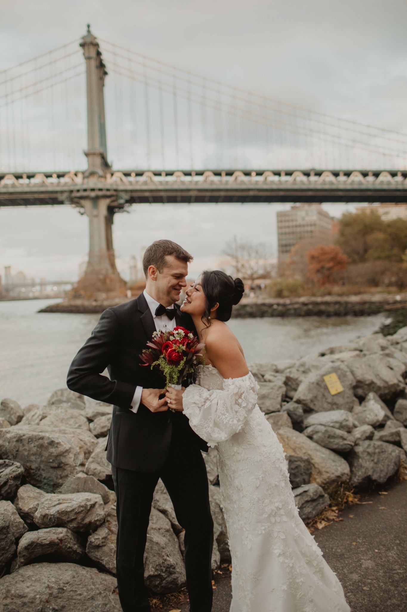 Couple in wedding attire standing on rocks near water with bridge in background, holding flowers and embracing.