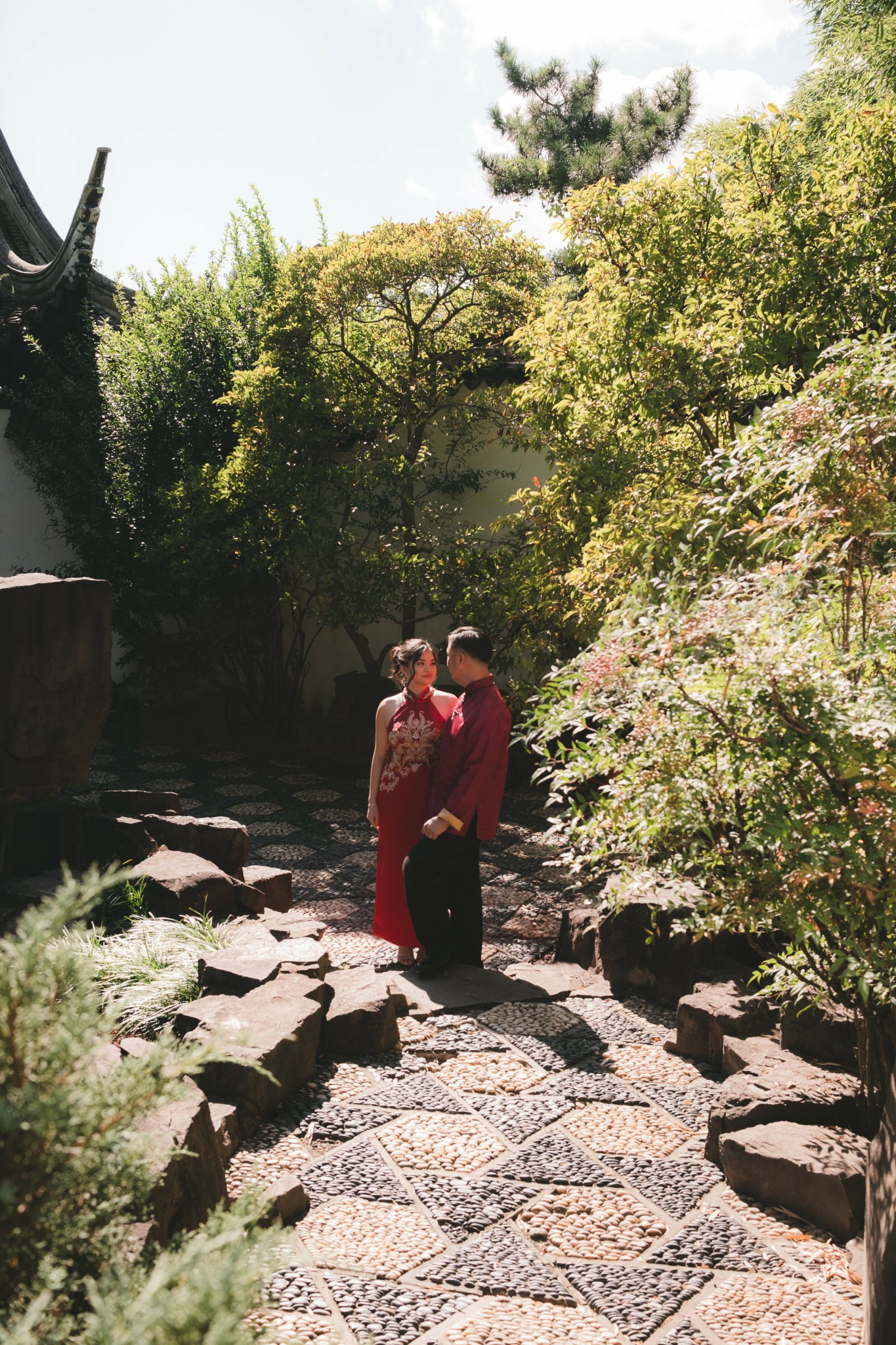 Couple standing on stone pathway surrounded by lush green trees and bushes in a garden.