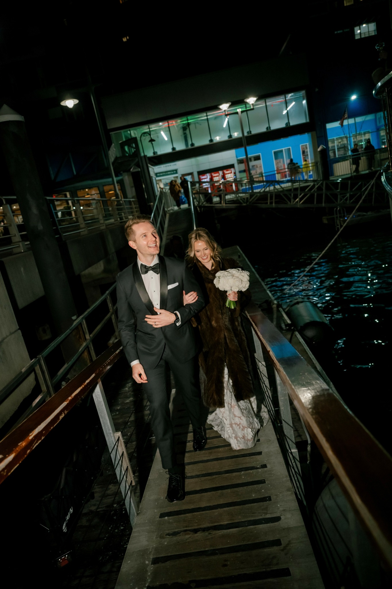 Two people in formal attire walking down a staircase at night near water, with buildings and lights in the background.