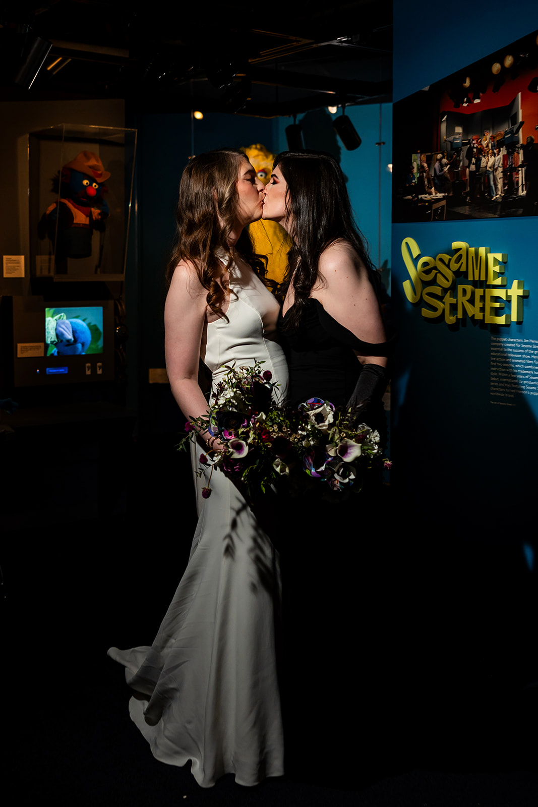 Two women in formal dresses kiss in a dimly lit room with a Sesame Street poster and a television screen in the background.