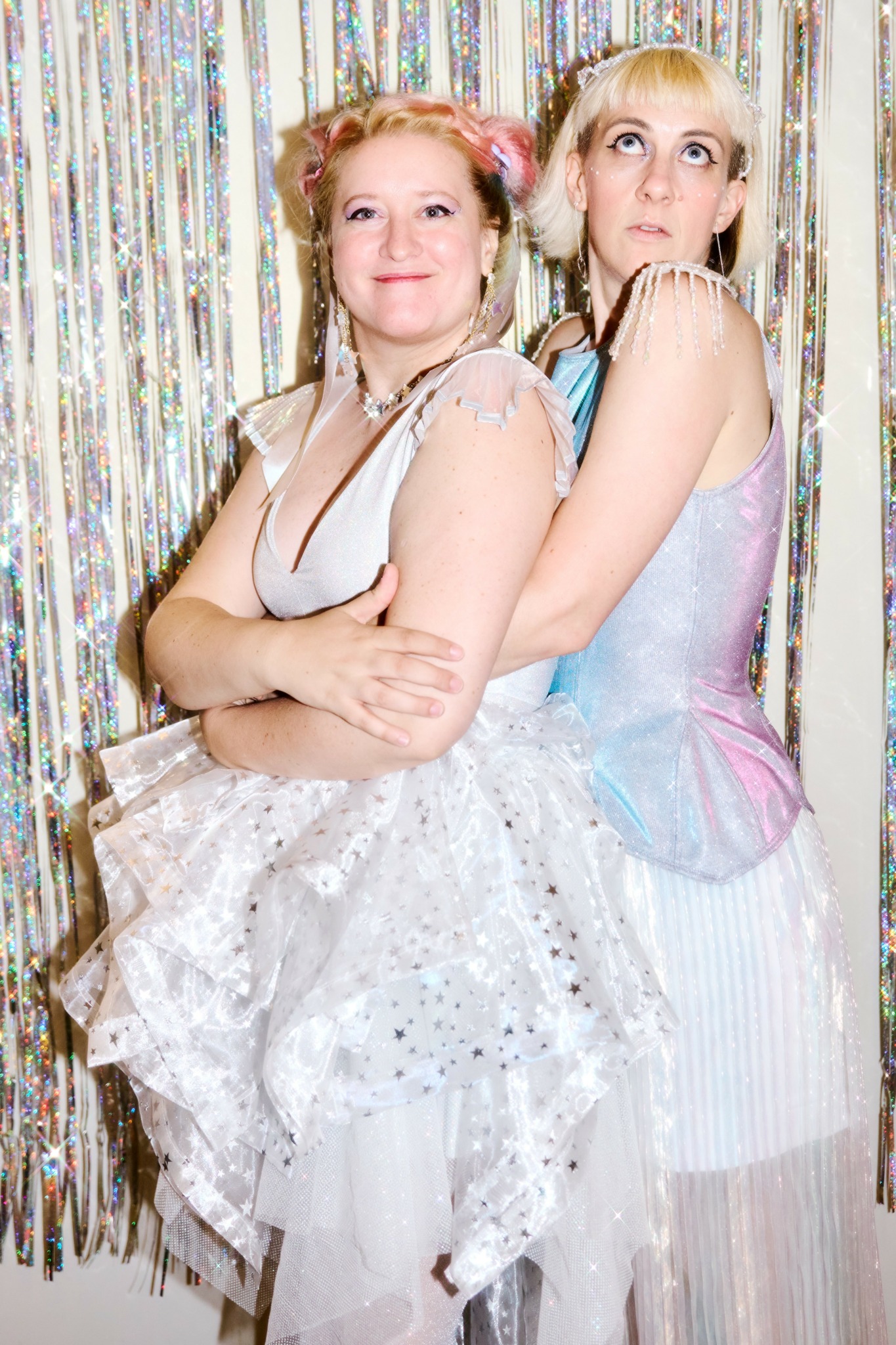 Two women in pastel dresses stand close together with arms crossed, smiling in front of a shiny, striped backdrop.