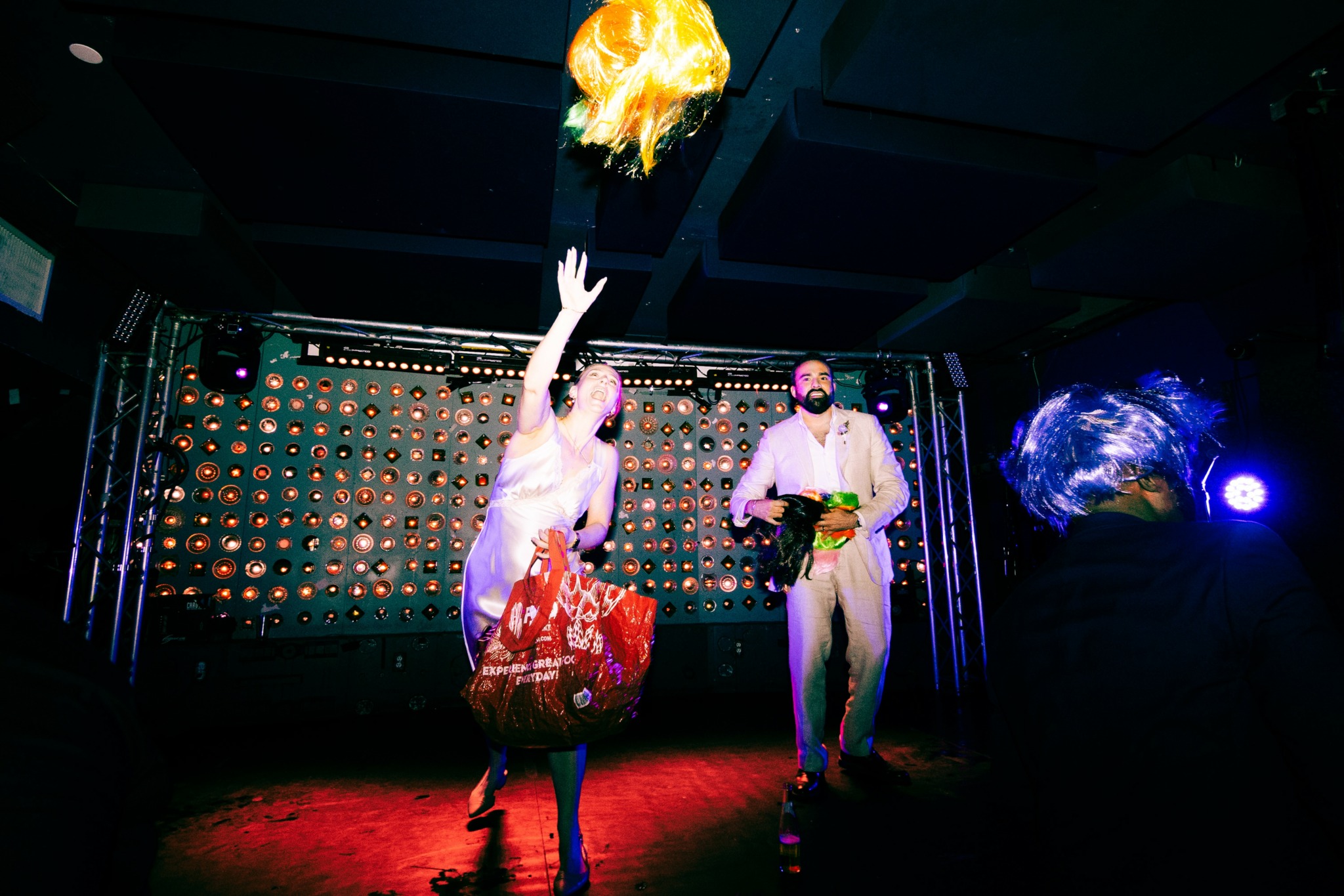 Person reaching for a flaming object above their head on a stage with two other people nearby, colorful background lighting.