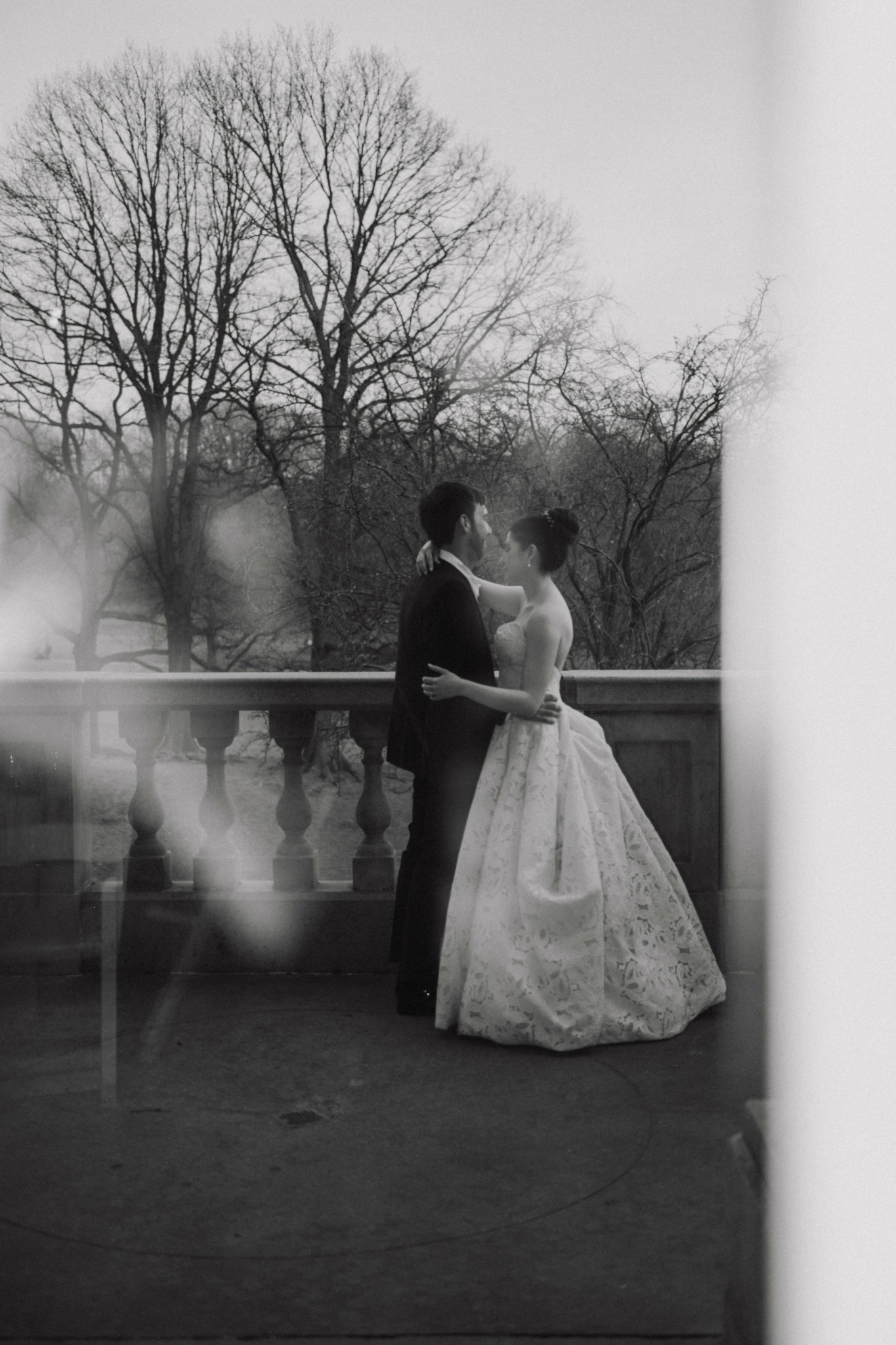 A couple in wedding attire standing on a balcony with trees in the background.