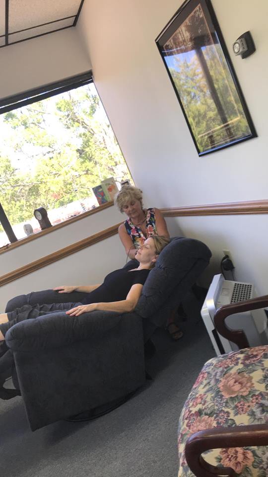 Person sitting in a recliner near a window with a woman standing behind them, in a room with framed picture and trees outside.