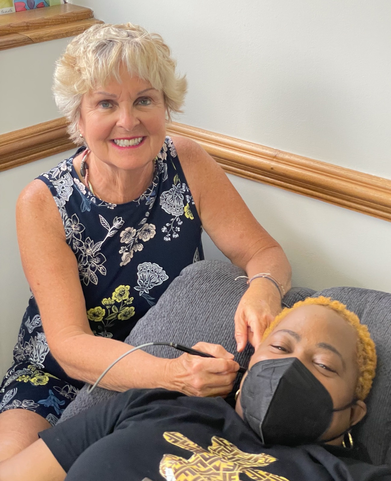 Woman smiling while holding a medical device near a person lying in bed wearing a face mask.