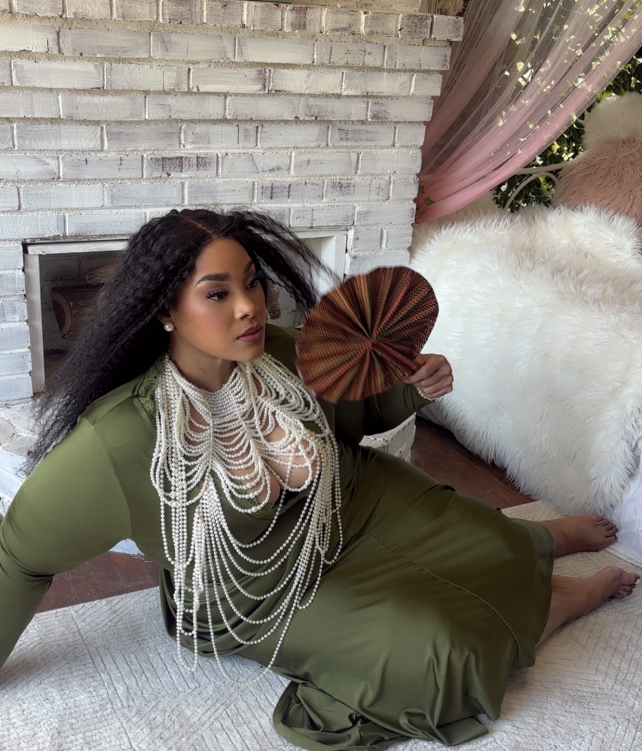 Woman with long curly hair sitting on a bed, holding a fan, in front of a white brick wall and pink curtains.