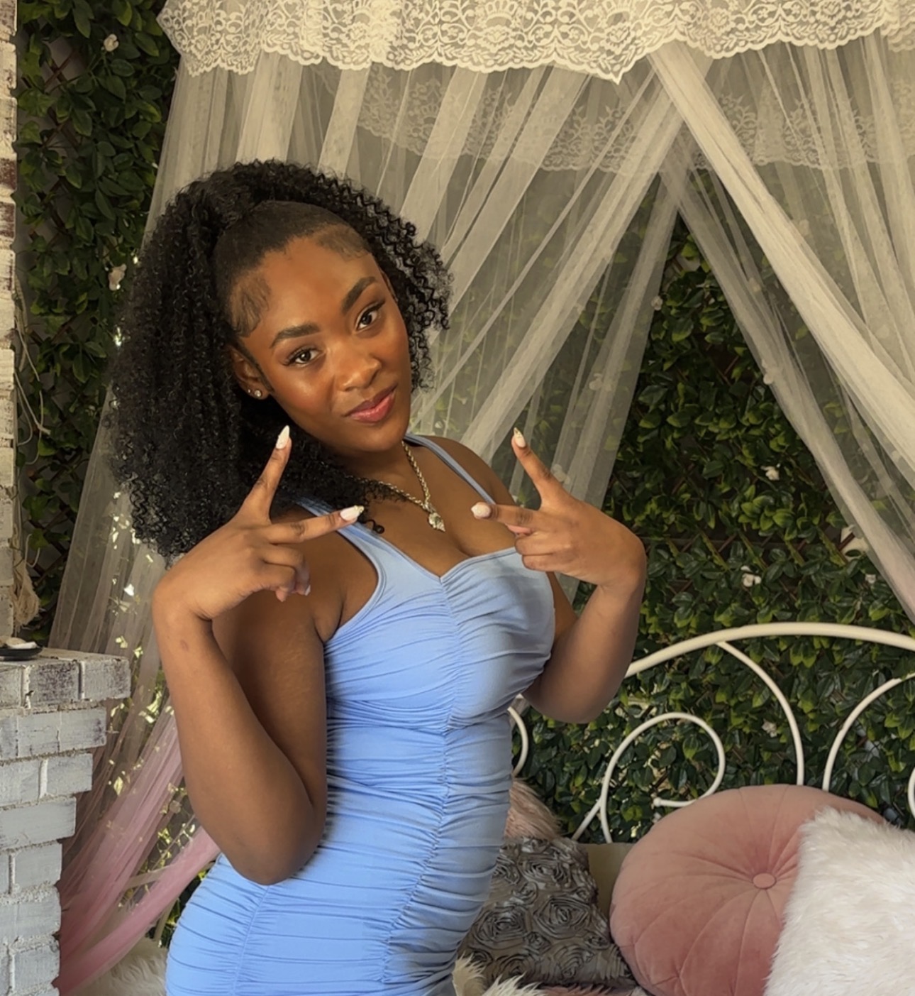 Young woman with curly hair in a light blue dress making peace signs with both hands, standing outdoors near a white metal bench.