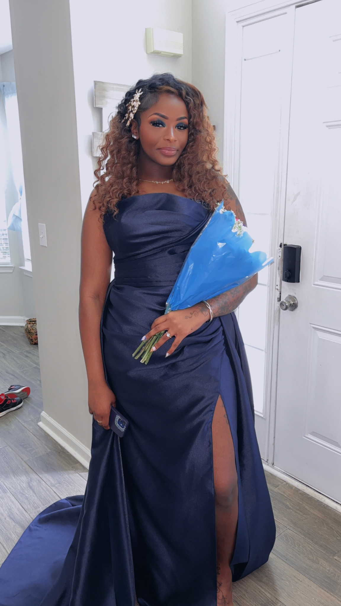 Woman in navy blue gown holding a bouquet, standing indoors near a white door and wall decor.