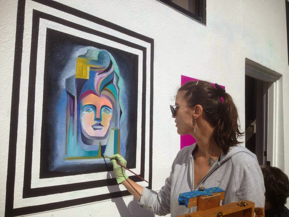 Woman painting a colorful abstract face on a large framed canvas in an indoor space.