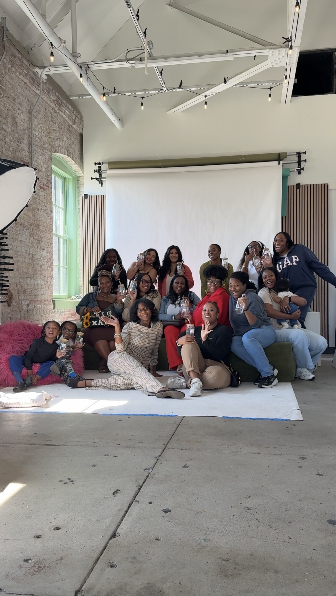 Group of people posing in front of a white backdrop inside a studio with brick wall and large window.