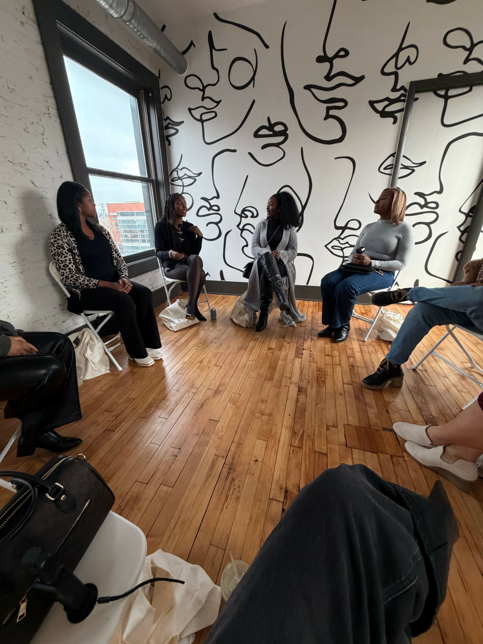 People sitting and standing in a room with wooden floor and a wall with abstract black line art.