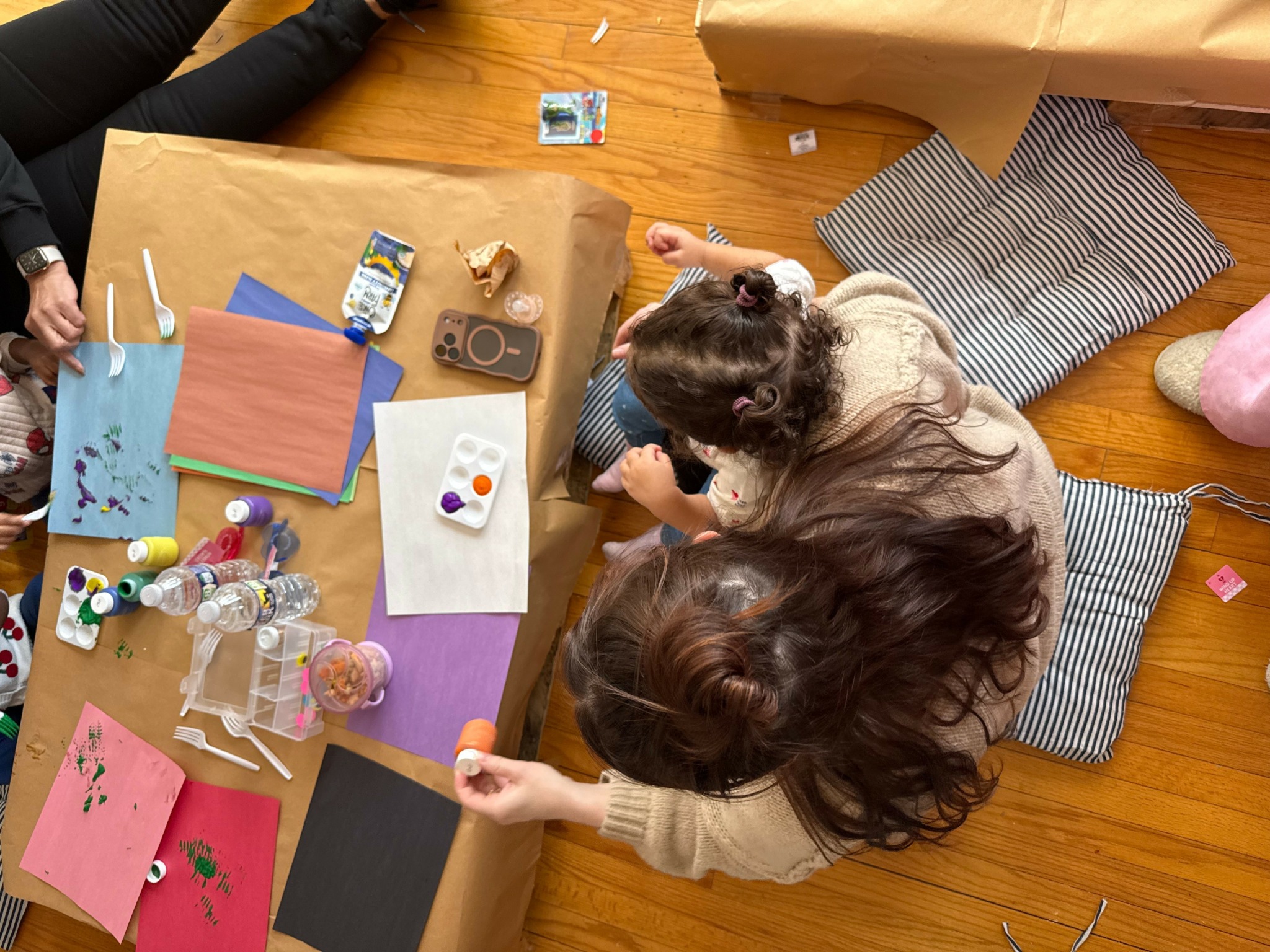 Two children sit at a table with craft supplies, wrapping paper, and pillows, working on a project together.