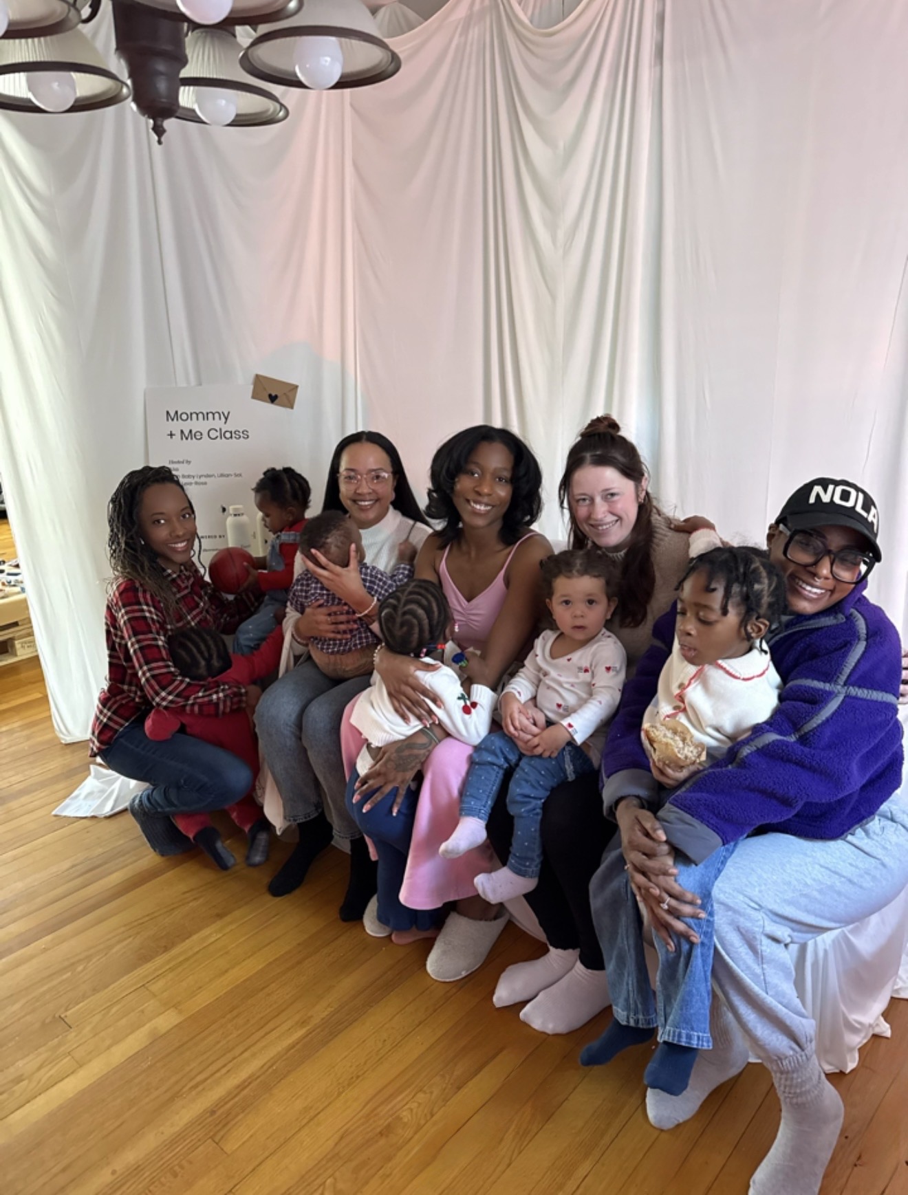 Group of women and children sitting on a couch in a room with white curtains and wooden floor.