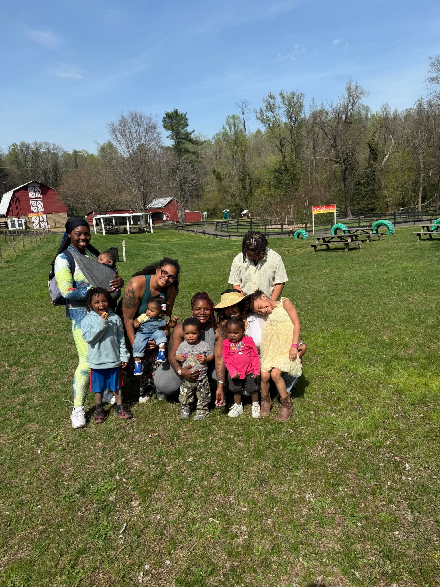 Group of children and adults standing on grass in a park with trees and playground equipment in the background.