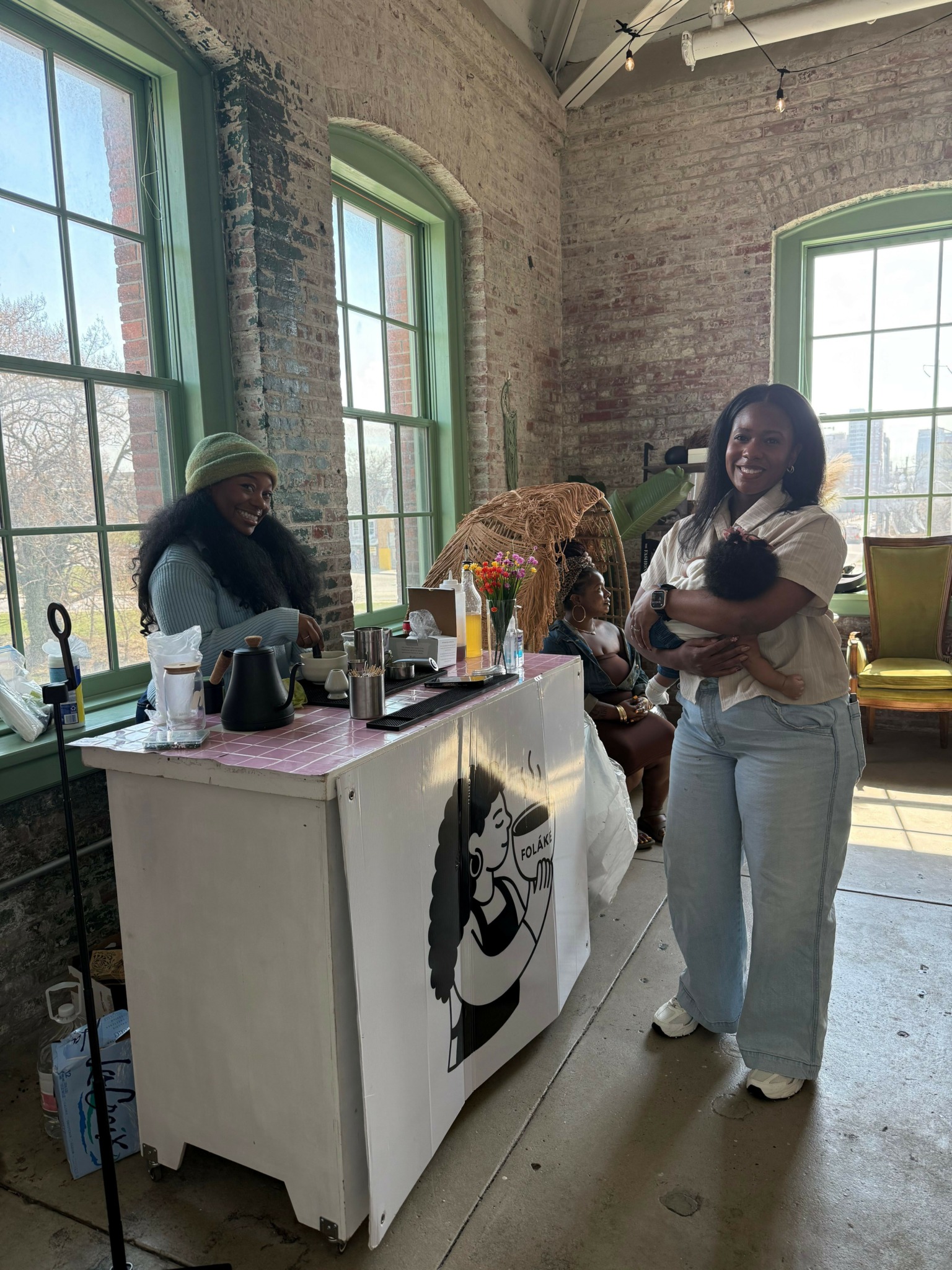 Two women and a dog near a table with art supplies in a room with brick walls and large windows.