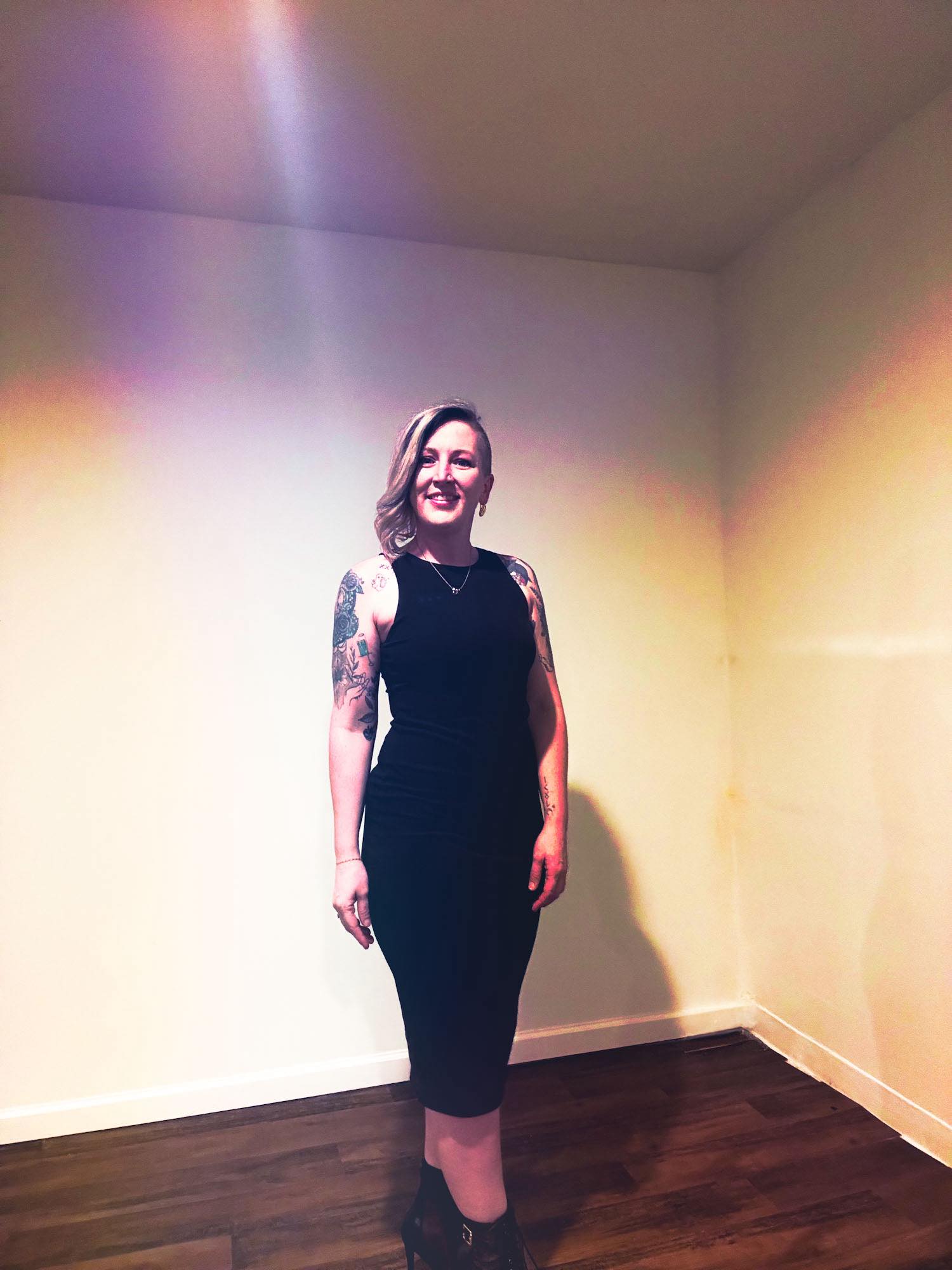 Woman with tattoos wearing a black dress standing in a room with plain walls and wooden floor.