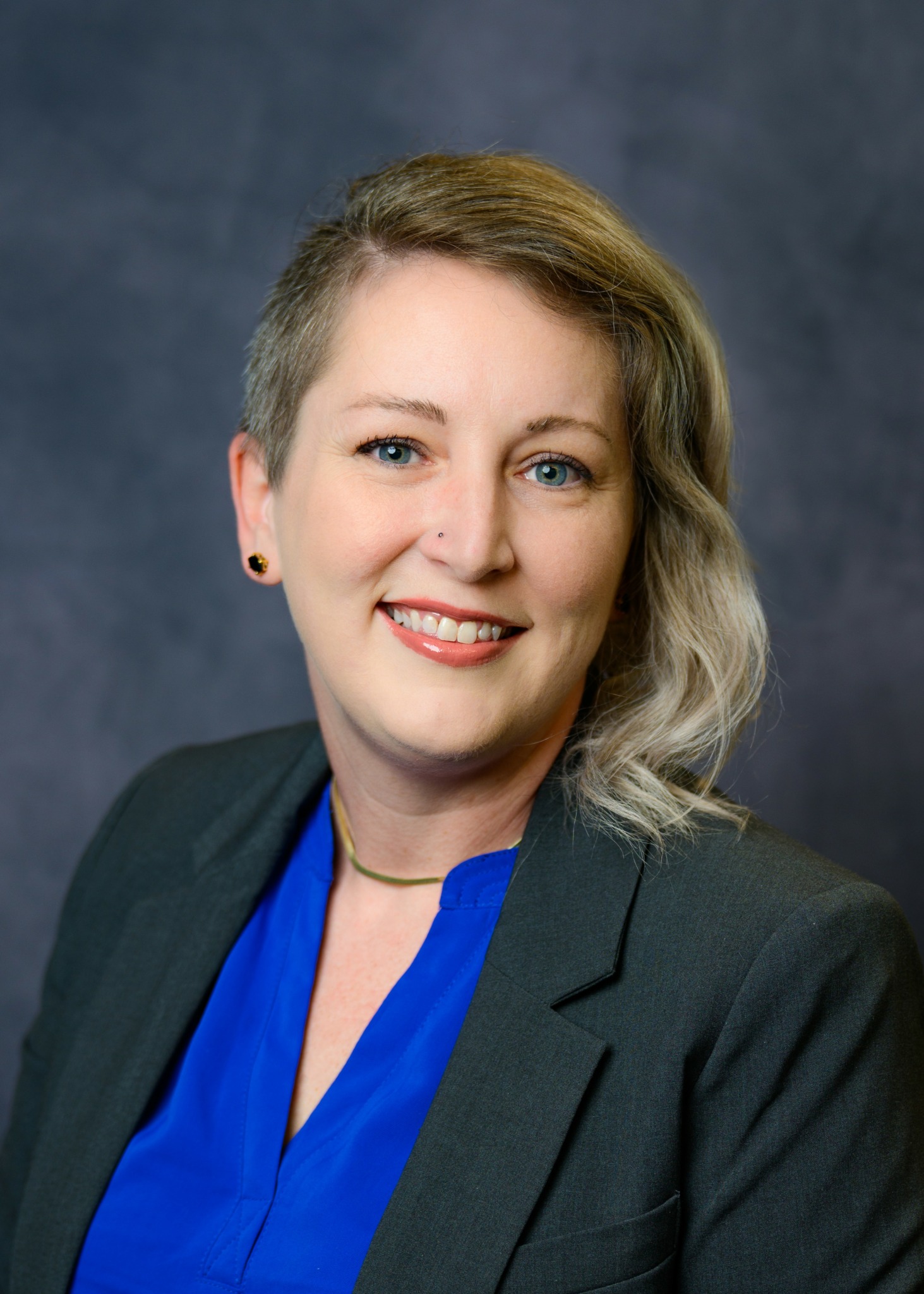 Portrait of a woman with short, wavy hair, wearing a dark blazer and blue blouse, smiling against a dark background.