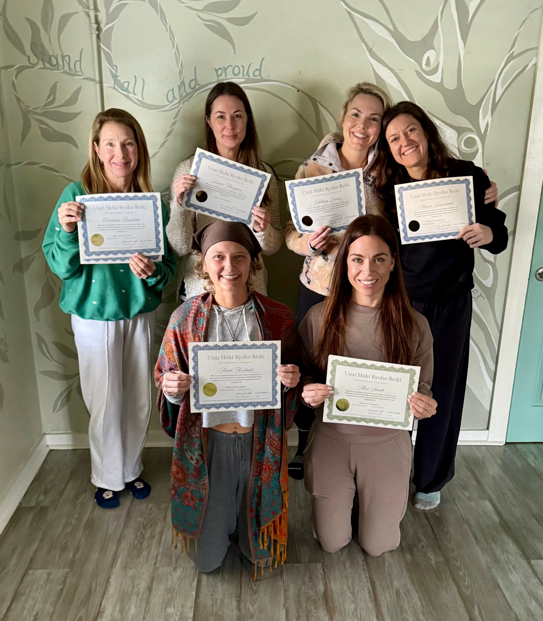 Six women holding certificates, standing in a room with a mural wall background, smiling at the camera.