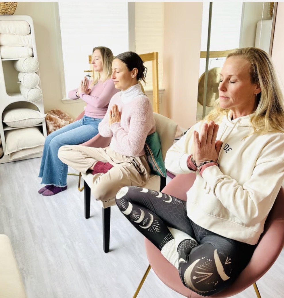 Three women sitting with eyes closed and hands in prayer position in a room with shelves and mirror.