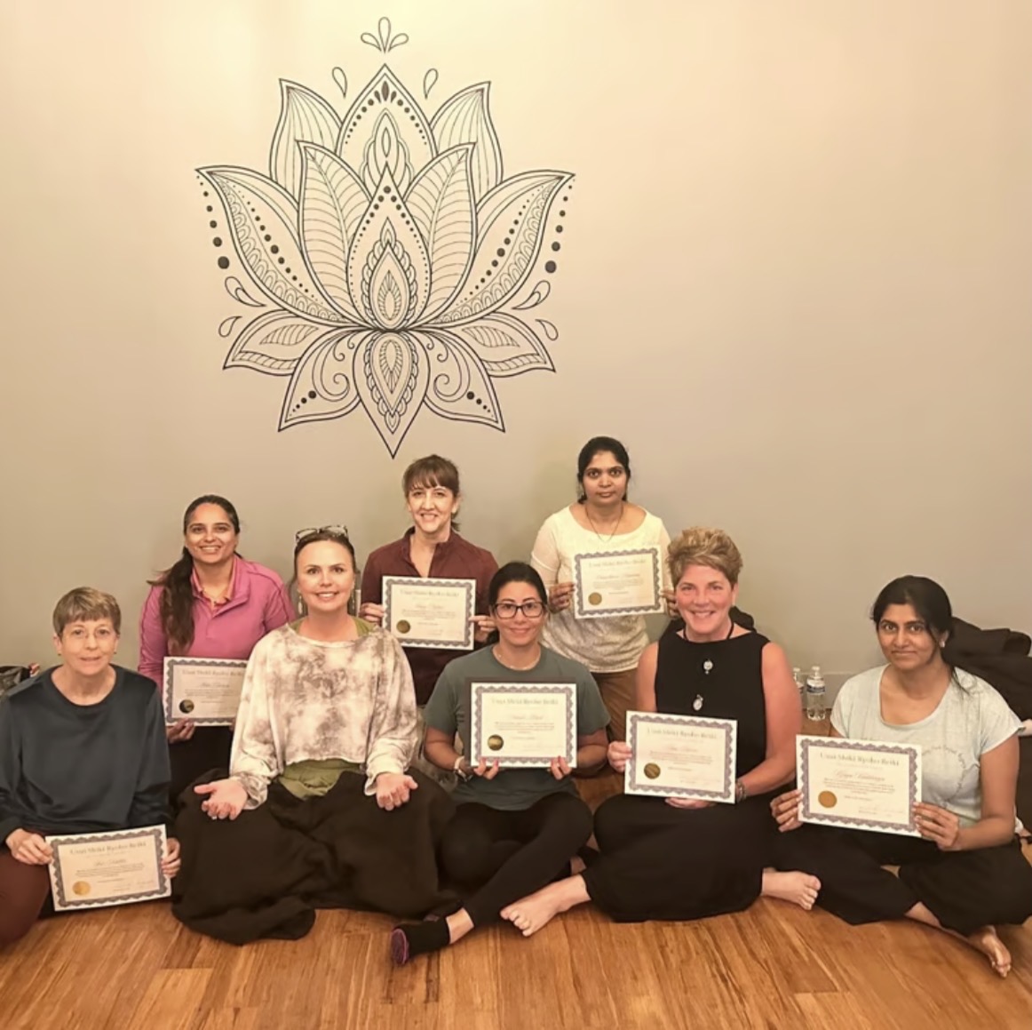 Seven people sitting and standing in front of a wall with a large lotus flower drawing, holding certificates.