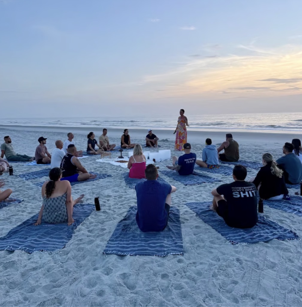 Group of people sitting on blue mats on sandy beach, listening to a woman speaking near the ocean at sunset.