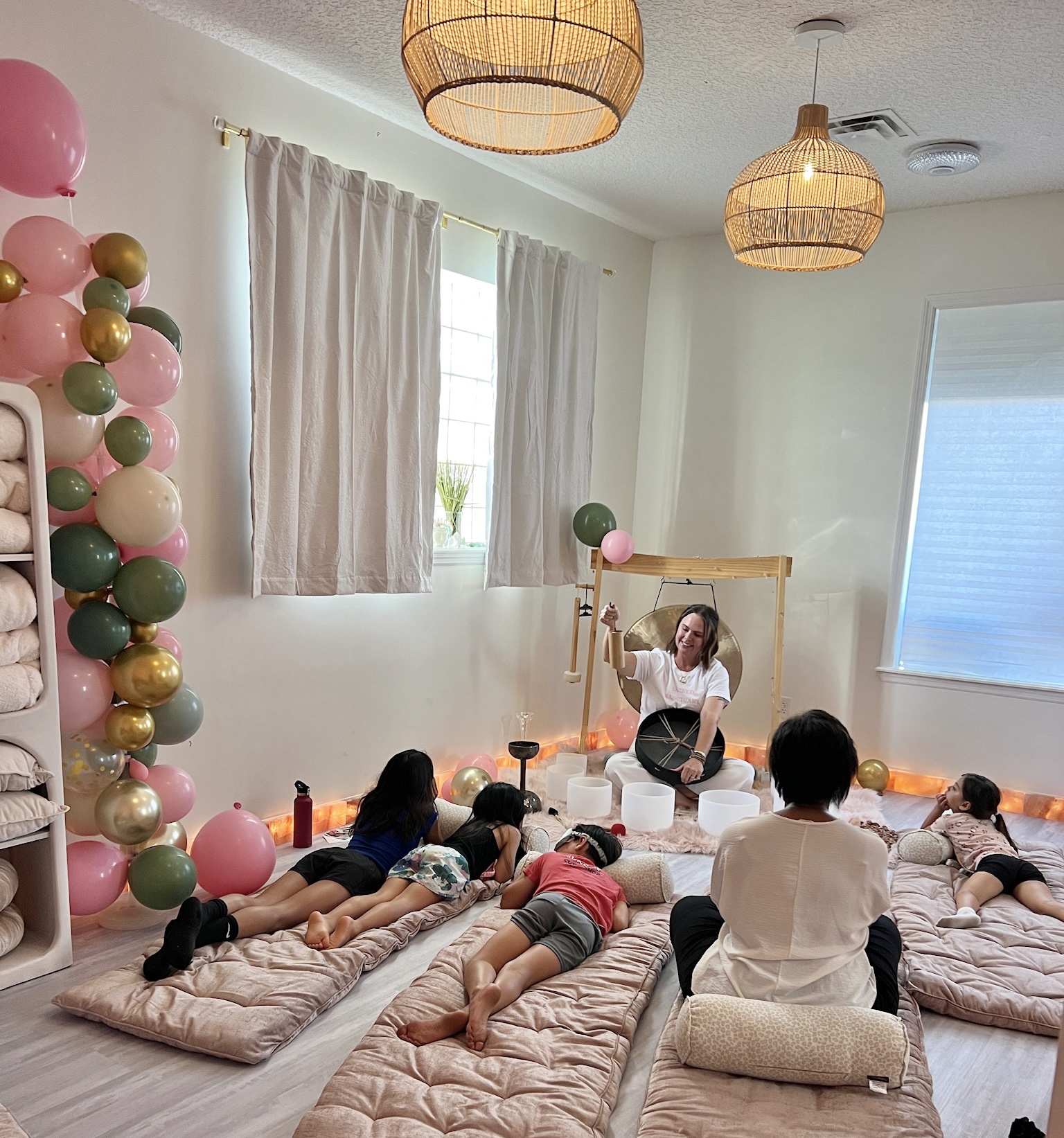 Children lying on mats during a yoga or relaxation session in a bright room with balloons and a woman leading.