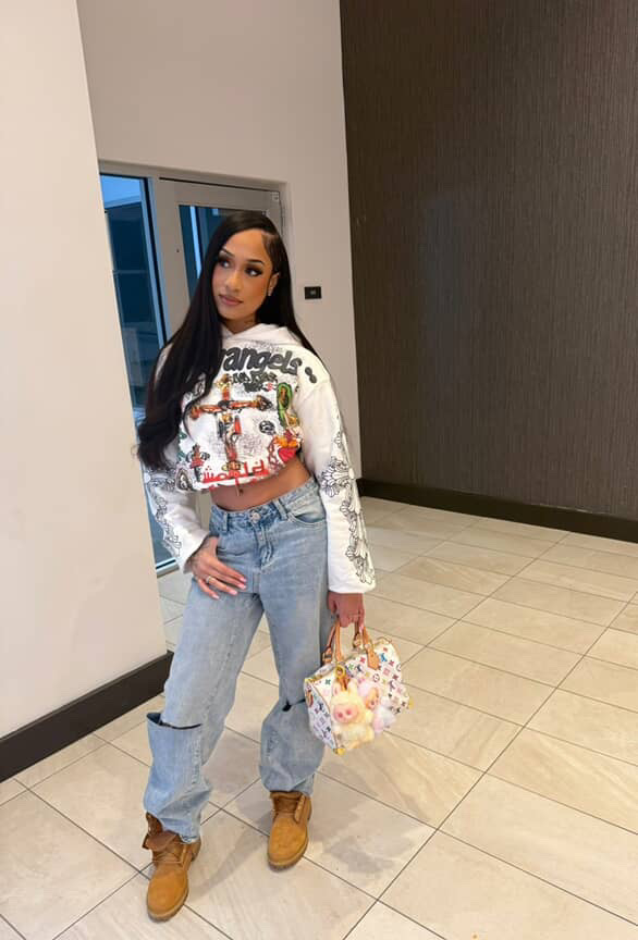 Young woman with long dark hair standing indoors, holding a floral handbag, wearing a colorful graphic crop top, jeans, and boots.