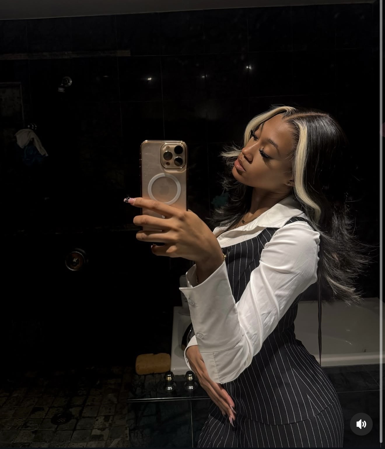 Woman with black and blonde hair taking a mirror selfie in a dark room, wearing a white shirt and pinstripe dress.