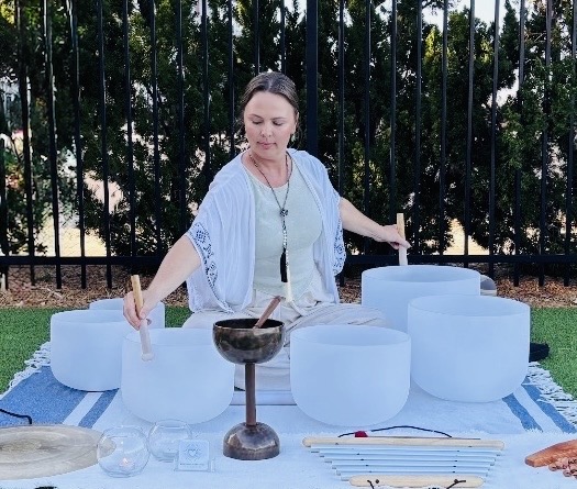 Woman playing crystal singing bowls outdoors with trees and a fence in background.