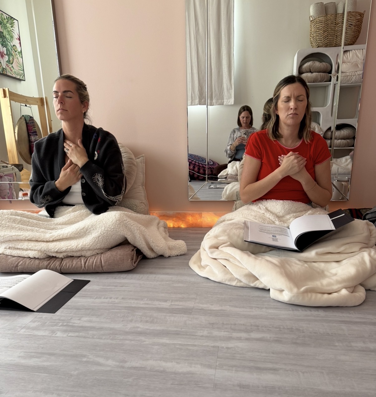 Two women sit on cushions with hands clasped, eyes closed, in a meditation or prayer session, in a cozy room.