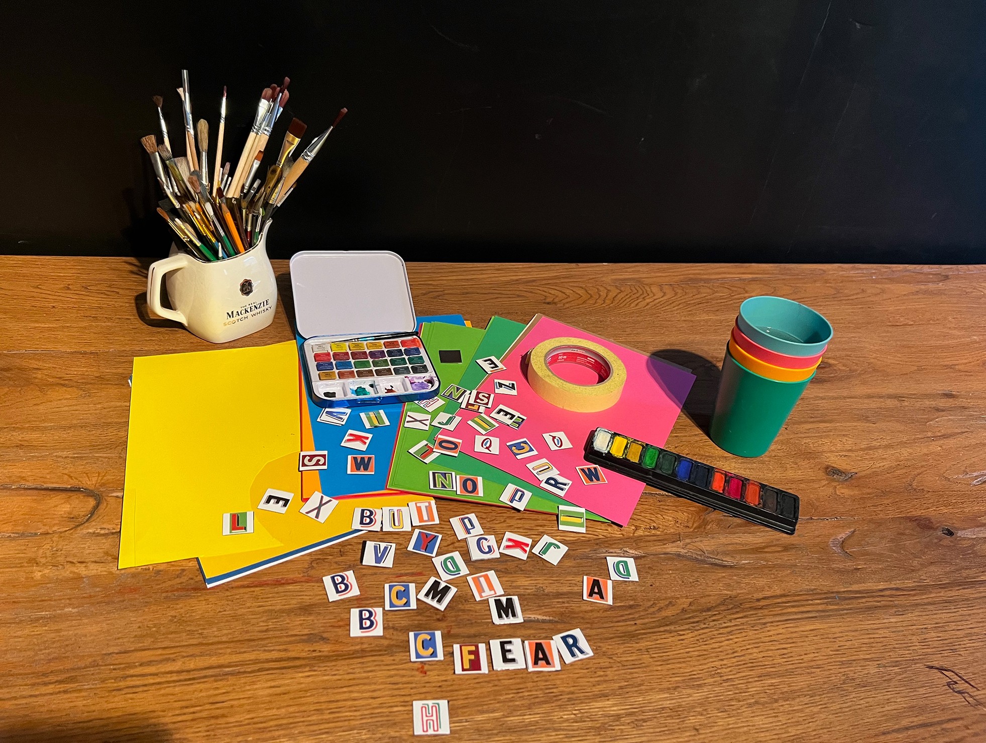 Art supplies and educational materials on a wooden surface, including paintbrushes, colored paper, tape, cups, and letter tiles.