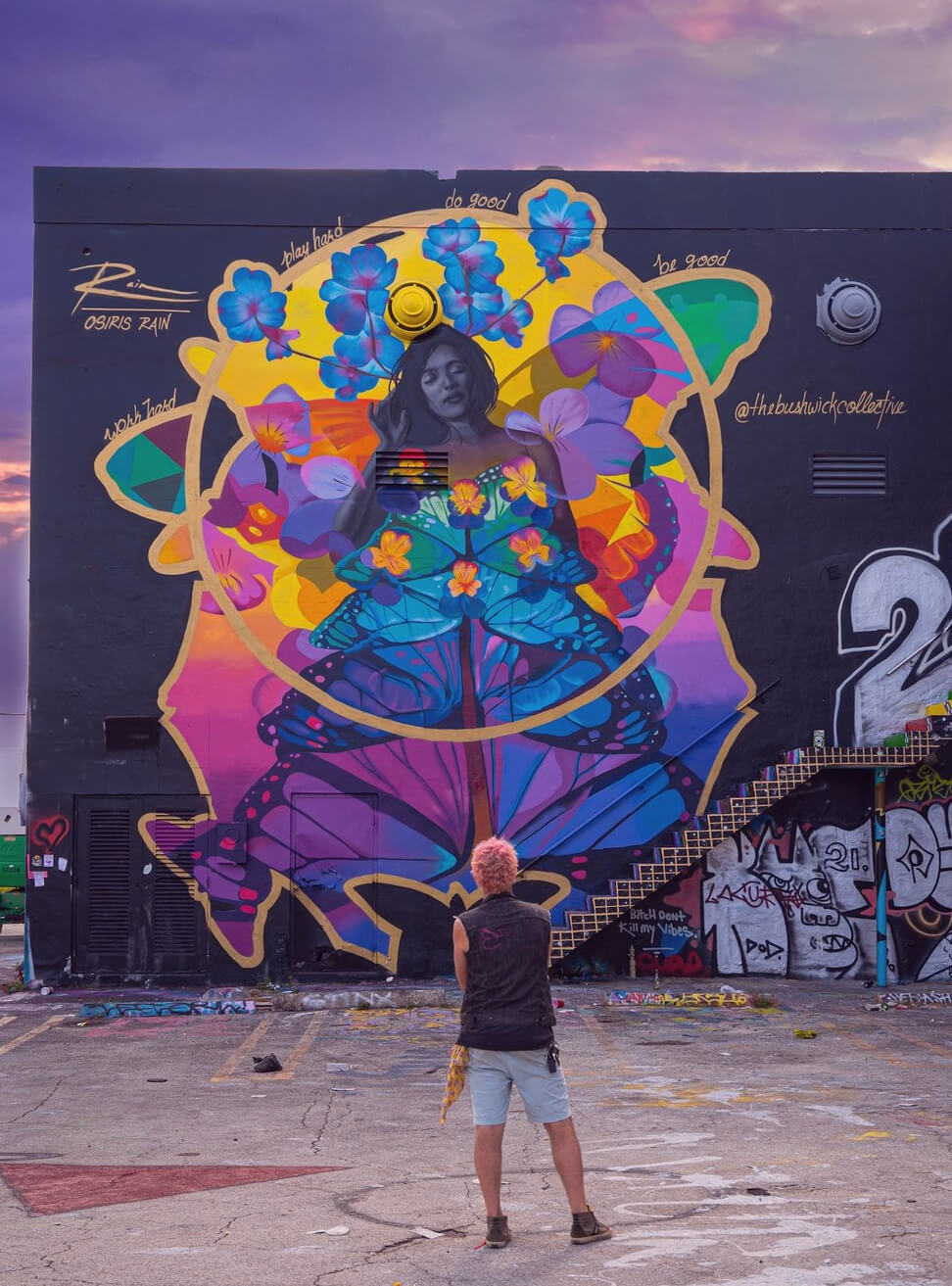 Person standing in front of colorful mural with a woman surrounded by flowers and abstract shapes.