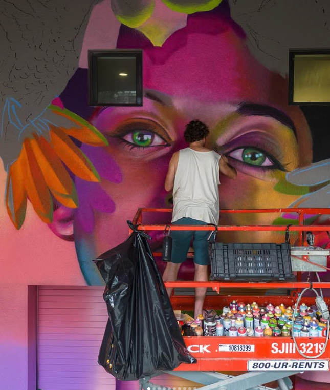 Person painting a colorful mural of a woman's face with green eyes on a wall.