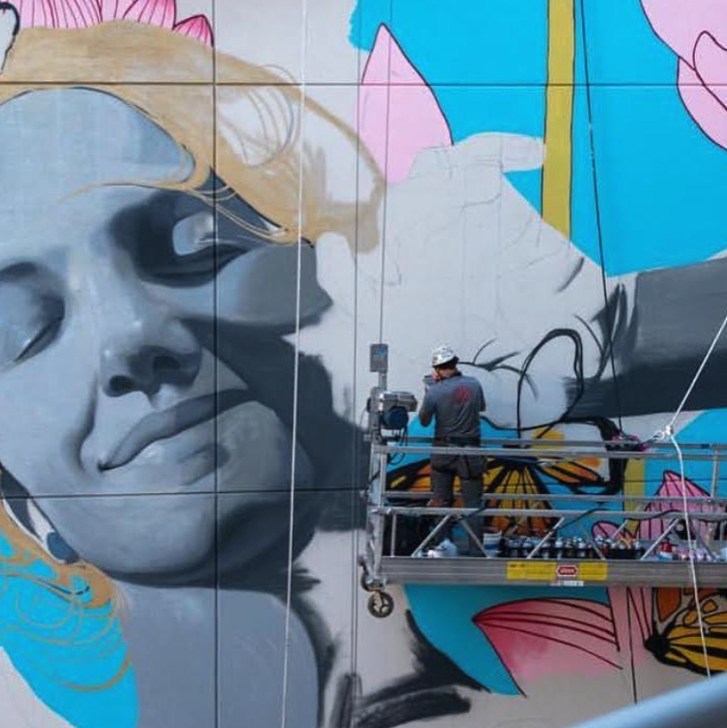 Artist working on a large mural of a woman's face with colorful background elements.