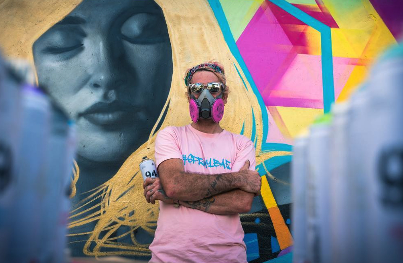 Man wearing a pink shirt and a mask, standing with arms crossed in front of a colorful mural with a face and geometric shapes.