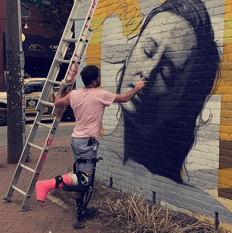 Person painting a large mural of a woman's face on a brick wall, using a ladder for access.