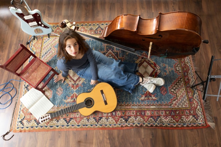 Woman sitting on a patterned rug with a guitar, surrounded by musical instruments and sheet music, looking up at the camera.