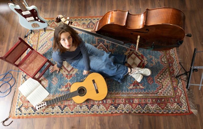 Woman sitting on a patterned rug with a guitar, surrounded by musical instruments and sheet music, looking up at the camera.