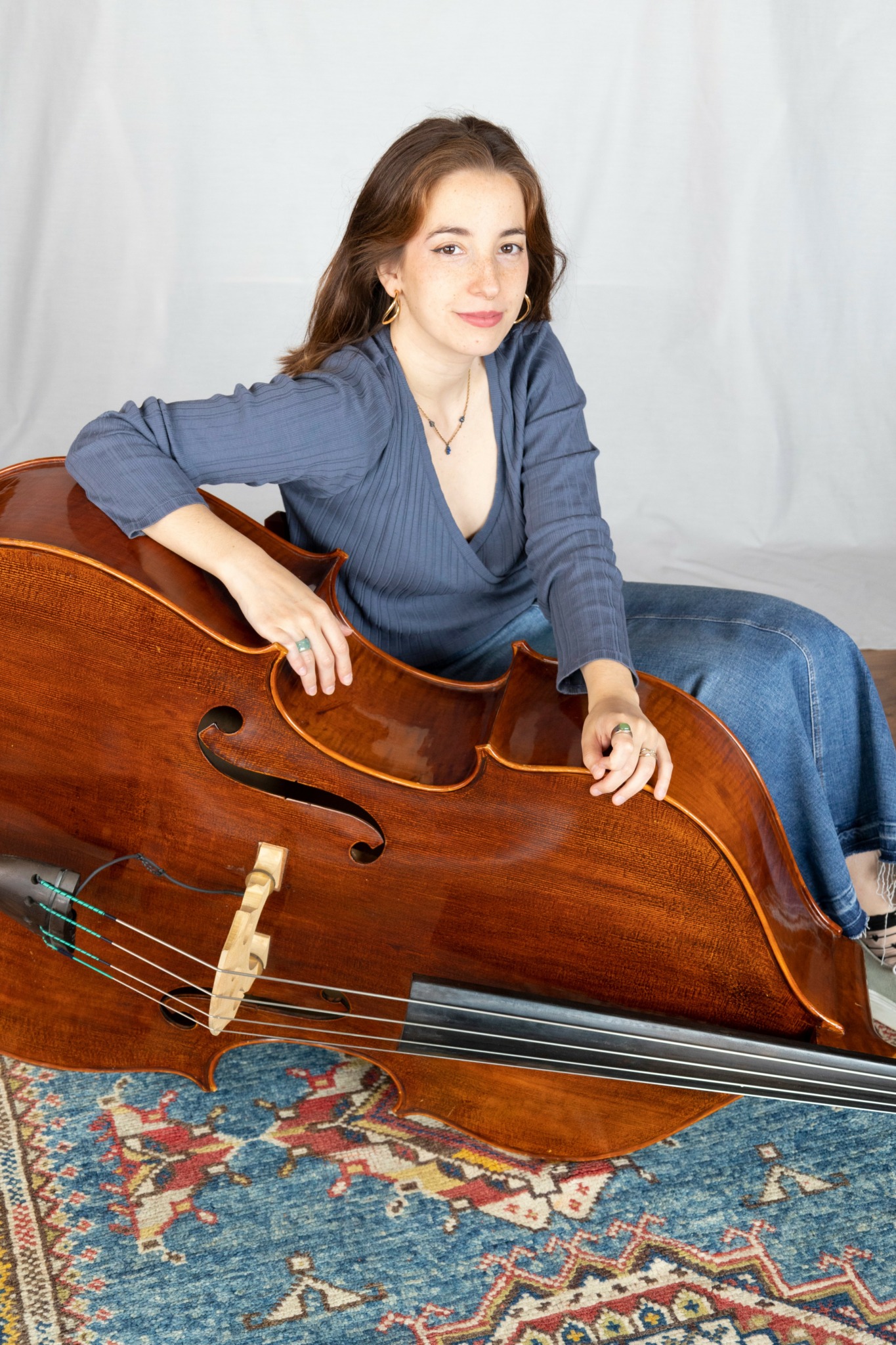 Woman with long brown hair sitting with a large wooden cello, wearing a blue top and jeans, on a patterned rug.